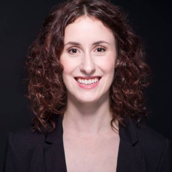 Alethea Bakogeorge, a white disabled woman with dark brown eyes and dark brown curly hair, smiles at the camera. She is wearing a black blazer and is standing against a black backdrop.