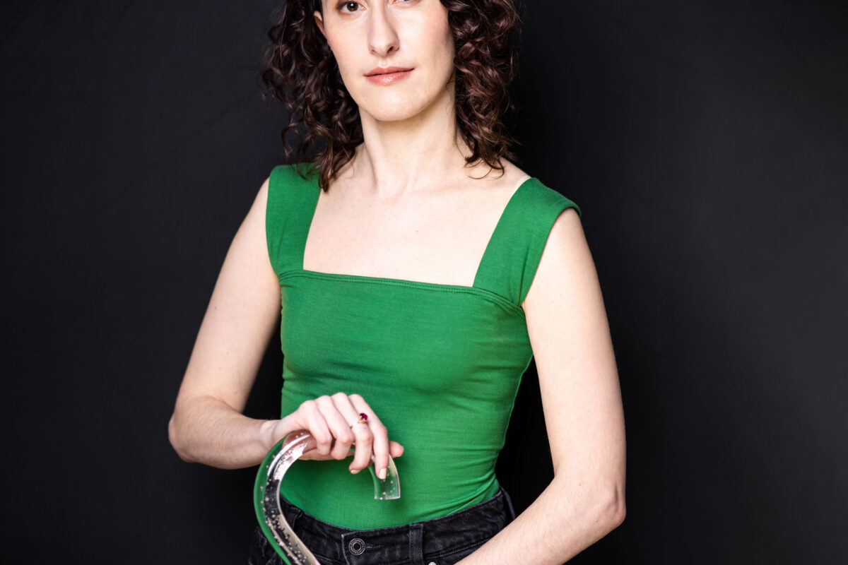 Alethea Bakogeorge, a white disabled woman with dark brown eyes and dark curly hair, poses for the camera with both hands on her lucite walking cane. She is wearing a kelly green tank top with wide straps and black jeans, and she stands in front of a black background.