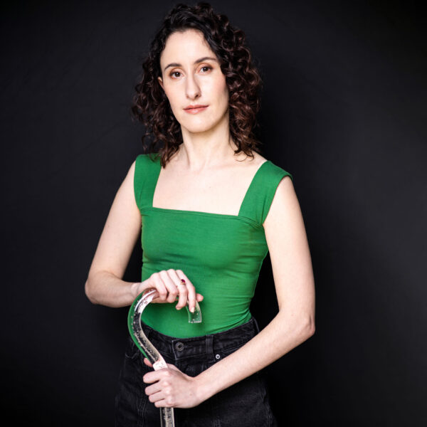Alethea Bakogeorge, a white disabled woman with dark brown eyes and dark curly hair, poses for the camera with both hands on her lucite walking cane. She is wearing a kelly green tank top with wide straps and black jeans, and she stands in front of a black background.