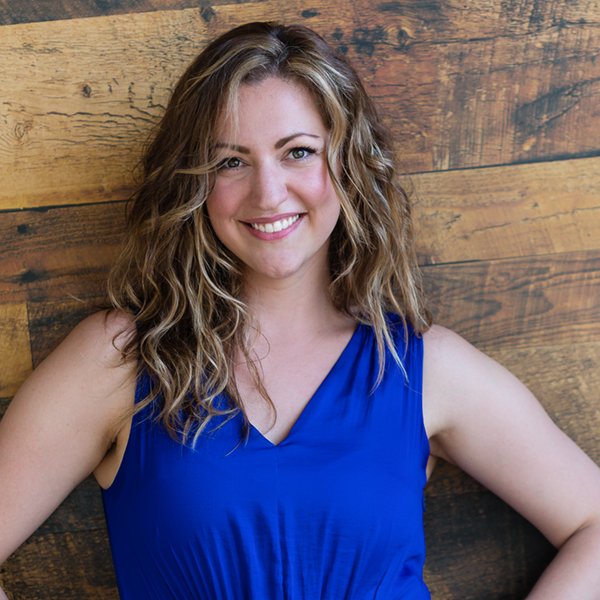 Kate Supleve, a white woman with long, light brown wavy hair and green eyes smiles at the camera. She is wearing a bright blue dress and stands in front of a wood-paneled wall with her hands resting on her hips.