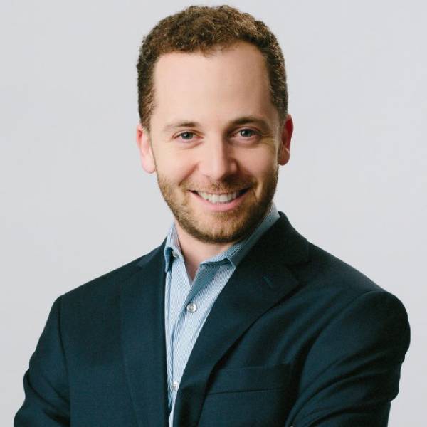 Mitchell Marcus, a white male with brown curly hair and a beard, smiles at the camera. He is wearing a blue sports jacket on top of a blue collared shirt.