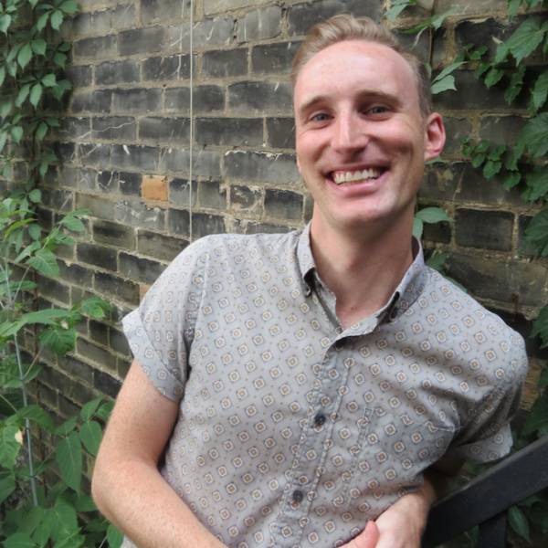 Paul Beauchamp, a white man with short light brown hair smiles at the camera. He is wearing a soft green patterned button-down short sleeves shirt, and is leaning up against an outdoor brick wall with hints of green foliage.