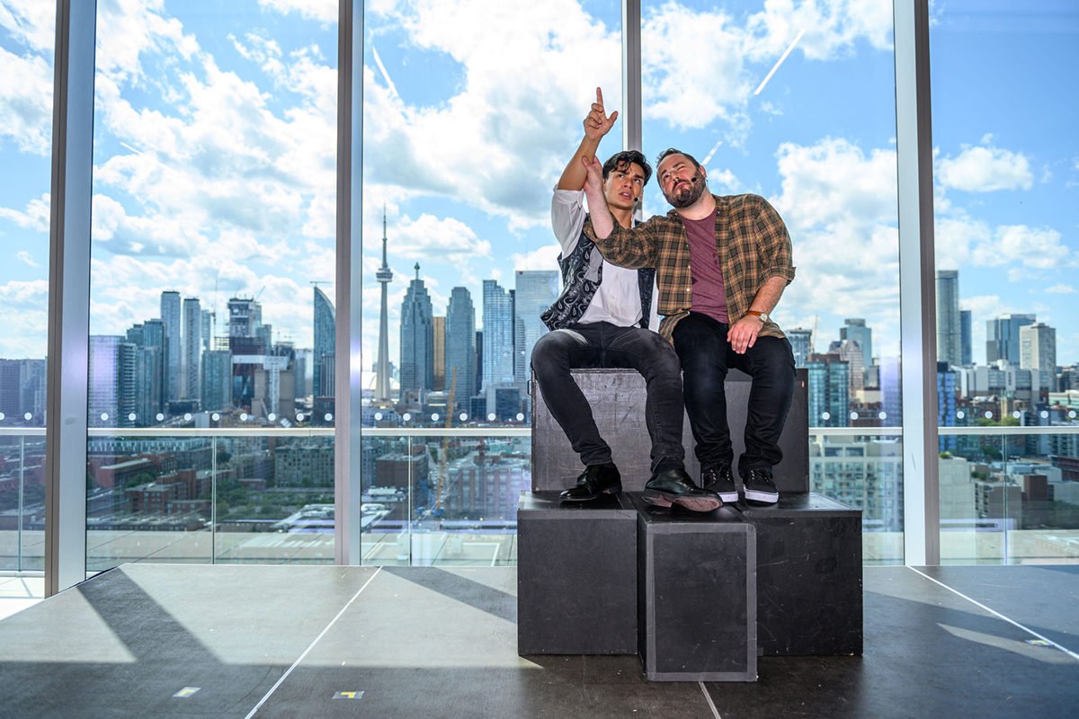 Two men sit huddled together on prop blocks, looking and gesturing at something high out of frame, one arm each, raised up.