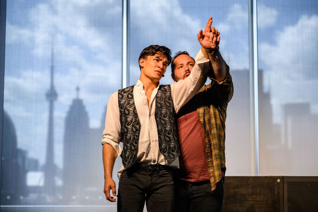 Two men are spotlighted on a stage, both staring up and off into the distance, close together and the one guiding the other's hand upward to point at a star.