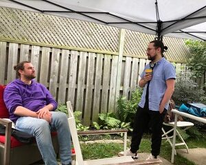 Bookwriter, Nick Green (left) is seated in a backyard chair, talking to music assistant, Jonathan Corkal-Astorga (right), who's standing while holding a drink. Nick is wearing a purple sweater, with his hair tied up in a bun, while Jonathan wears a blue button-up & bun.
