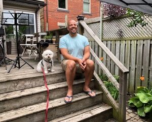 Deputy Artistic Director, Ray Hog (right) sitting on backyard patio steps, smiling while seated next to Bobby the dog, the musical mascot (left). He is wearing a blue tee & shorts, while Bobby is a small white fluffy dog, on a red leash.