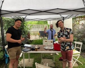 Composer, Kevin Wong (left), music assistant, Jonathan Corkal-Astorga (middle), & music supervisor, Chris Barillaro (right), are all standing around a backyard patio table, underneath a pop-up awning. They are all smiling at the camera and in summer attire.
