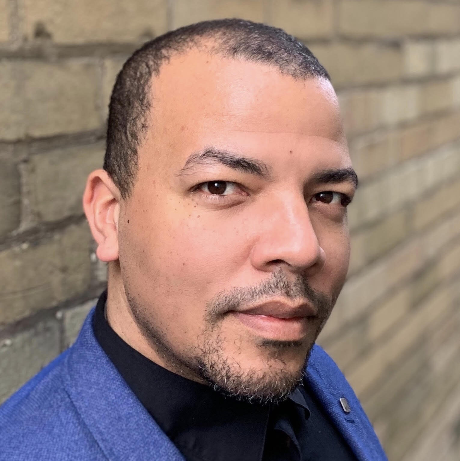 Ray Hogg, a Black man, stands in three quarter profile looking directly into the camera with a slight smile and warmth in his eyes. He has close cropped balck hair, a widow's peak dark eyebrows and a goatee. He is wearing a blue blazer, a black dress shirt and stands against a yellow brick background.