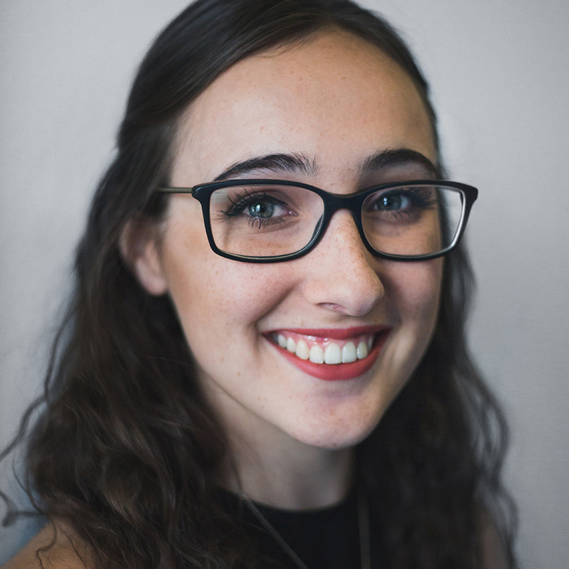 Cassandra Di Felice is a white woman with glasses, blue eyes, and dark brown hair. She is smiling at the camera, wearing a black high-neck top