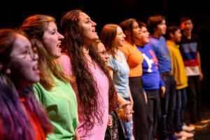 Multiple youths of various backgrounds, lined up on stage and singing as a chorus