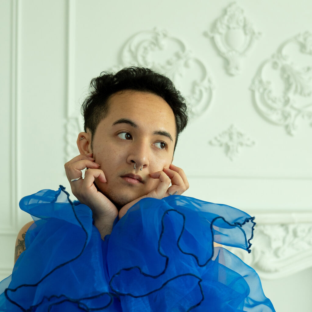 Cameron Vindua is a mixed race genderqueer person, with light brown skin. He has short curly brown hair and dark brown eyes. They're posed in large ruffled sleeves, while looking off-camera.