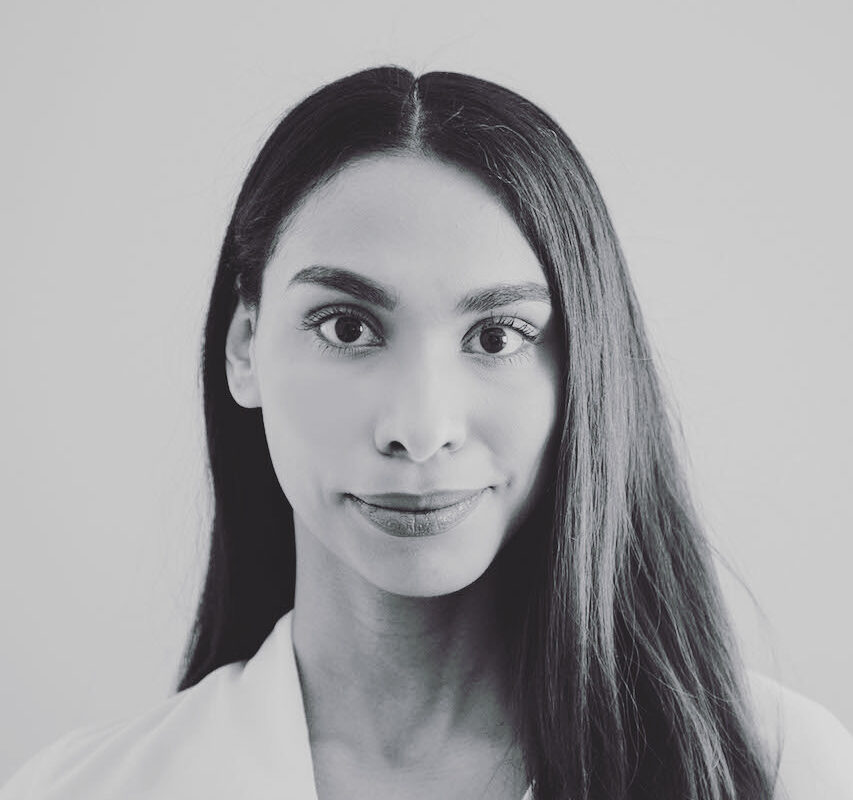 Patricia Cerra is a person of colour who is smiling at the camera in a black and white photo. She is wearing a white shirt with her long dark hair parted down the middle and falling over her left shoulder.