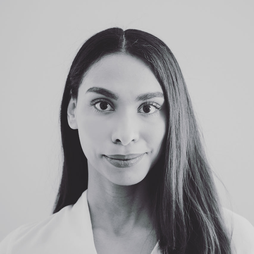 Patricia Cerra is a person of colour who is smiling at the camera in a black and white photo. She is wearing a white shirt with her long dark hair parted down the middle and falling over her left shoulder.