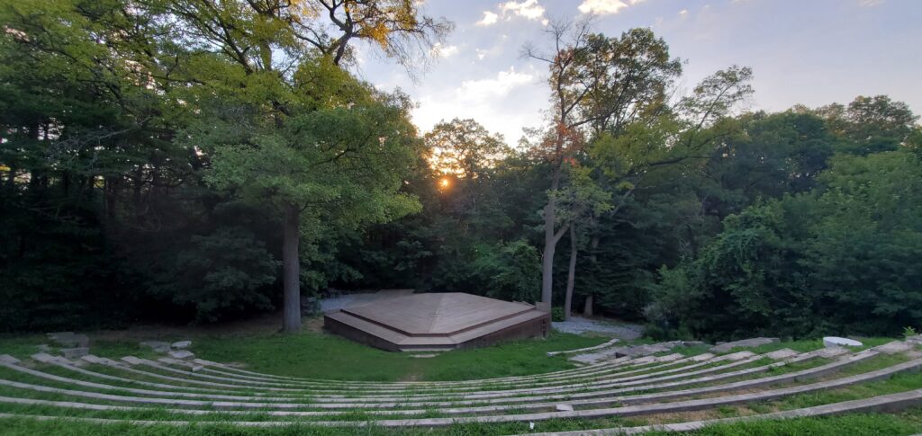 An image of long winding stone step/seats curved in a forest clearing, centering a large wooden square stage, surrounded by trees. There's small fluffy clouds and a sunset. 
