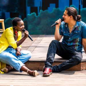 Two performers are on an outdoor wooden stage, seated close together and looking at each other as they sing, a painted backdrop in the background. Germaine (left), a dark-skinned Black Femme, is seated on the lower-stage step, their legs slightly crossed as they lean forward, singing into the mic they hold. Dillan (right), a young Indigenous Ojibwe performer with long, dark hair is seated on the lower-stage step, leaning back and facing Germaine, smiling as he sings into his mic.