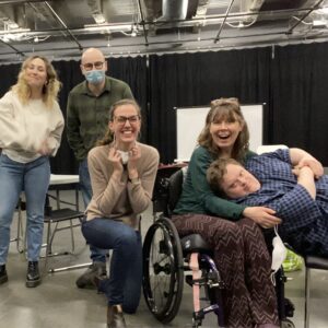 5 white people are posed together in a theatre auditorium, all smiling at the camera. Two stand toward the back of the group (one masked), with a third person kneeling in the center. A fourth person is seated in a wheelchair and hugging/pillowing someone to their right, with the fifth person leaning into the embrace across their chair. Pictured left to right: Lucy McNulty, Anton Lipovetsky, Ali Joy Richardson, Debbie Patterson, Niall McNeil.