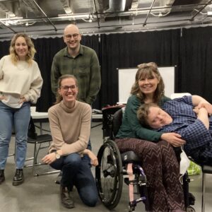 5 white people are posed together in a theatre auditorium, all smiling at the camera. Two stand toward the back of the group, with a third person kneeling in the center. A fourth person is seated in a wheelchair and hugging/pillowing someone to their right, with the fifth person leaning into the embrace across their chair. Pictured left to right: Lucy McNulty, Anton Lipovetsky, Ali Joy Richardson, Debbie Patterson, Niall McNeil.