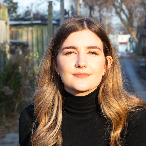 Katie is a queer, caucasian person, in her mid-twenties. She has blondish/brownish hair and hazel eyes under dark eyebrows. She is wearing a black turtleneck, and her hair is half-tucked behind her right ear. She is standing in an alley with garages and trees, that are just slightly out of focus. She is staring just past the camera and half-smiling while being lit by the glow of the sun.