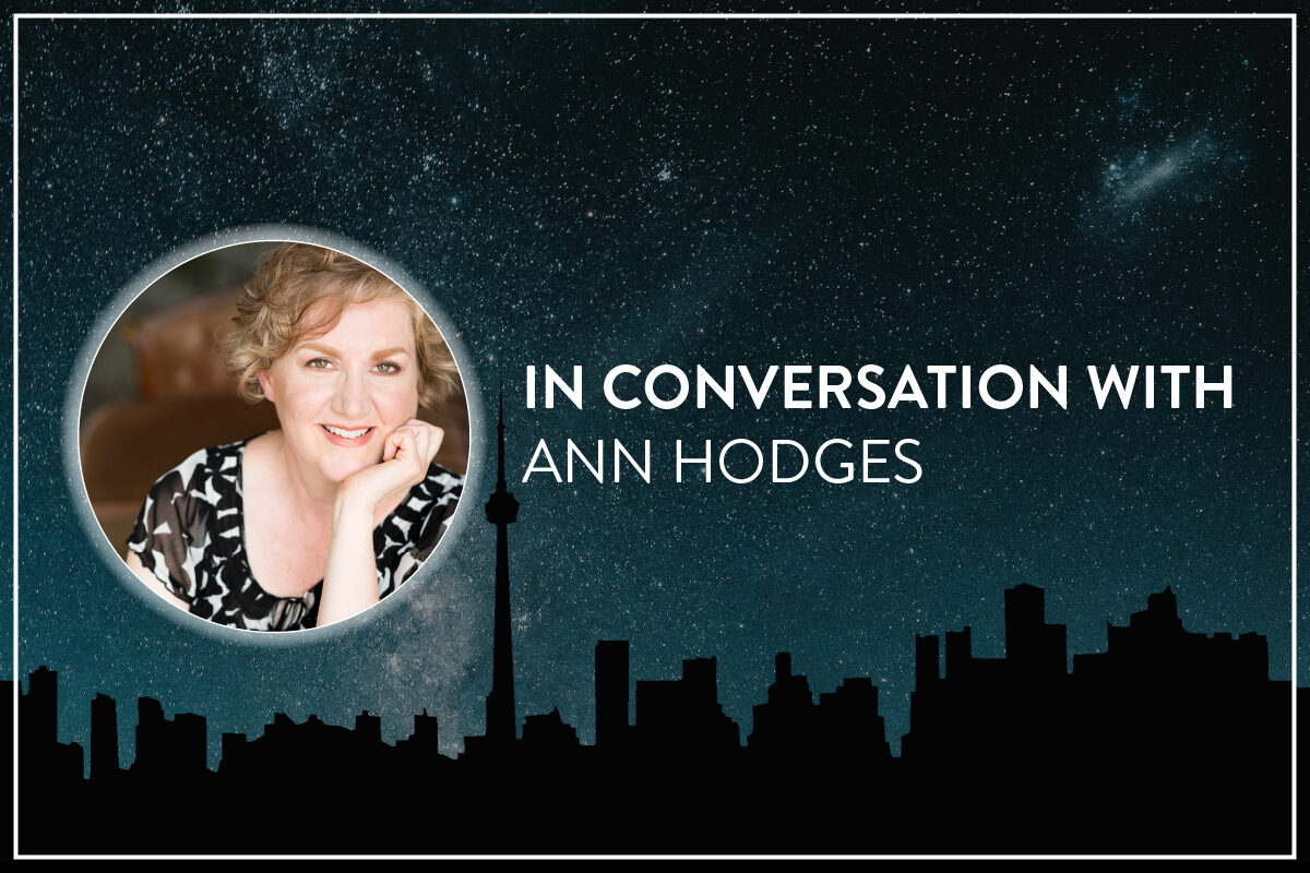A background of the Toronto skyline silhouette runs along the bottom, against a blue, star filled galaxy sky. Centered is a circular headshot and text. The headshot is a smiling white woman (Ann Hodges), with a white glow around the circle edge. Bold white text to the right of the image reads, "In conversation with / Ann Hodges."