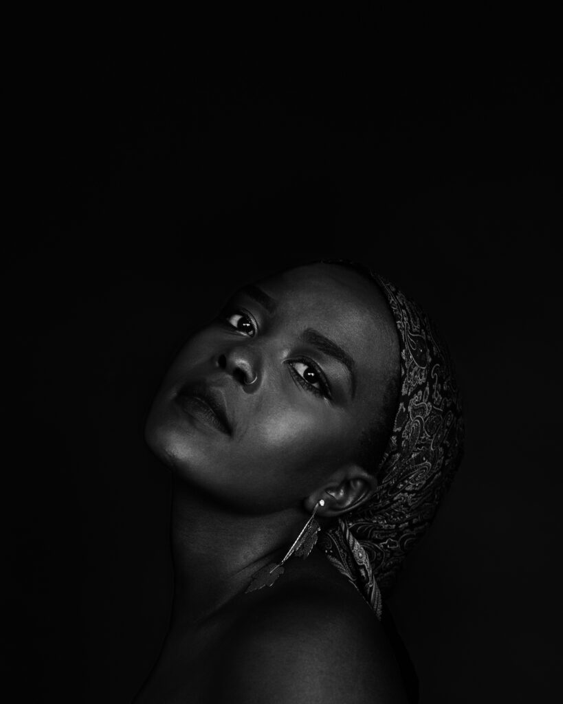 A black and white photo of Germaine Konji, a dark skinned Black Femme in her mid twenties with warm brown eyes and black hair wrapped in a patterned headscarf. She’s looking meaningfully at the camera, dignified against the black background with her head tilted back.