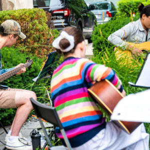 An angled image of 3 musicians seated at a distance from each other and each playing a different type of guitar. They have music stands and sheet music in front of their seat, all surrounded by greenery in a hedged in garden patio.