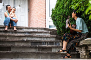 An image of an outdoor front porch with several stone steps leading up to a white door. An Asian parent (left) is seated on the top step with a young toddler in their lap, smiling at Vicky Wang (right). Vicky is seated at the bottom of the steps on a concrete bench, smiling up at her audience, as she plays guitar amongst shrubbery.