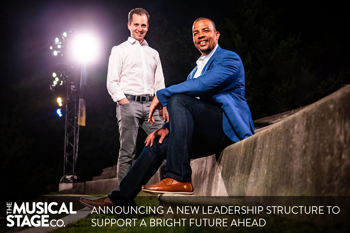 Two men are posed outdoors on tall concrete amphitheatre seats at night, with bright outdoor overhead lighting. Mitchell (left) is a white man with brown curly hair, standing and smiling at the camera. He is wearing a white dress shirt and grey pants. Ray (right) a Black man with close cropped black hair, a widow's peak, dark eyebrows and a goatee, sits centre, with one knee bent, smiling at the camera. He is wearing a blue blazer, white shirt and dark wash jeans. White text along the bottom reads, “Announcing a new leadership structure to support a bright future ahead.” The Musical Stage Company logo in white sits to the left of the text.