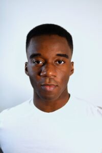 Matthew Joseph (he/they), a black non-binary artist with dark brown eyes and a fade, delivers a warm but subtle smirk to the camera. He is wearing a white crew neck shirt and standing against an eggshell white background.