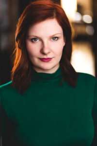 Rosie Callaghan, a white woman with red shoulder-length hair, looks at the camera with a small smile. She wears a green turtleneck.