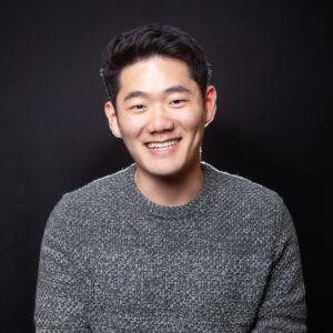 Haneul is a Korean-Canadian performer. In this image, he is wearing a knitted gray sweater, and is smiling facing the camera.