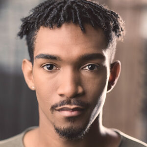 David Andrew Reid is a medium-brown skinned male of African-Jamaican descent. He has black hair styled in twists on the crown of his head and faded sides, dark brown eyes looking directly at the camera, a black moustache and chin beard. He is wearing an olive-green round-neck shirt