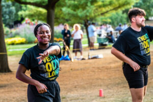 A shot of two instructors in matching t-shirts and black shorts outside at a park - mid dance move as they lead a dance class. Left, a Black femme instructor with a mini head-mic, smiles at the camera, their left arm reaching across their body. Right, a white masc instructor looks out of frame as they dance. Park goers mill in the background.