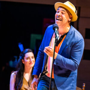 An angled image of Beau Dixon, a Black man with warm eyes and a moustache, wearing a blue blazer and light trilby hat, brightly lit on stage and singing into a standing mic while smiling at someone out of frame. He’s posed as if mid-stride and mid-blink. Two performers are seated in the background, smiling.
