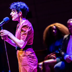 A side profile image of Sara Farb, a white woman with short, brown curly hair, wearing a belted, clay-colour jumpsuit, spotlighted on stage and singing into a standing mic. Her eyes are closed mid-note, her hands wrapped around the stand as she leans forward. Two other performers are seated in low blue lighting in the background.