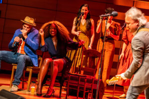 A full-body image of Beau Dixon and Jully Black, seated on stage and partly highlighted while they dance in their seats. Beau (left), a Black man with warm eyes and a moustache, wearing a blue blazer and light trilby hat, plays harmonica into the mic he’s holding; and Jully (right), a Black woman of Jamaican-Canadian descent with long, full earth-toned hair and blonde highlights, wearing a belted navy wrap dress, leans into Beau, her eyes closed and smiling brightly in mid-dance. Other performers are standing behind them or out of focus in the right frame.