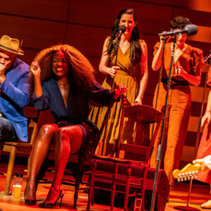 A full-body image of Beau Dixon and Jully Black, seated on stage and partly highlighted while they dance in their seats. Beau (left), a Black man with warm eyes and a moustache, wearing a blue blazer and light trilby hat, plays harmonica into the mic he’s holding; and Jully (right), a Black woman of Jamaican-Canadian descent with long, full earth-toned hair and blonde highlights, wearing a belted navy wrap dress, leans into Beau, her eyes closed and smiling brightly in mid-dance. Other performers are standing behind them or out of focus in the right frame.