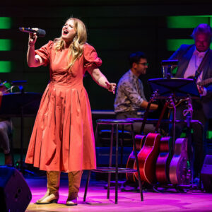 Kelly Holiff, a white woman with long blonde hair and green eyes, wearing an orange southern belle dress and cowboy boots, stands spotlighted on on stage, holding up her mic as she sings, eyes closed in the moment. The orchestra is low-lit behind her in dark blues and pinks, stripes of green lighting framing the shot.