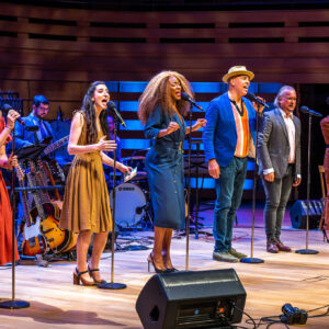 An angled distanced image of 6 performers lined up and spotlighted on stage, as they sing into their standing mic. Left to right shown are Kelly Holiff, a white woman with long blonde hair and green eyes, wearing an orange southern belle dress and cowboy boots; Hailey Gillis, a white woman with long wavy brown hair half-tucked back, wearing a mustard cocktail dress; Jully Black, a Black woman of Jamaican-Canadian descent with long, full earth-toned hair and blonde highlights, wearing a belted navy wrap dress; Beau Dixon, a Black man with warm eyes and a moustache, wearing a blue blazer and light trilby hat; Andrew Penner, a white man with brown and silver hair and a grey blazer, & Sara Farb, a white woman with short, brown curly hair, wearing a belted, clay-colour jumpsuit. Behind them are glimpses of the orchestra and several instruments.
