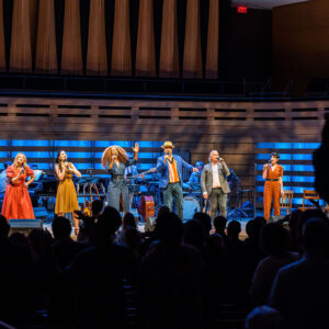 A centered, distanced image of 6 performers lined up and spotlighted on stage, as they sing into their mic and encourage the standing audience to clap along and dance. Left to right shown are Kelly Holiff, a while woman with long blonde hair and green eyes, wearing an orange southern belle dress and cowboy boots; Hailey Gillis, a while woman with long wavy brown hair half-tucked back, wearing a mustard cocktail dress; Jully Black, a Black woman of Jamaican-Canadian descent with long, full earth-toned hair and blonde highlights, wearing a belted navy wrap dress; Beau Dixon, a Black man with warm eyes and a moustache, wearing a blue blazer and light trilby hat; Andrew Penner, a white man with brown and silver hair and a grey blazer, & Sara Farb, a white woman with short, brown curly hair, wearing a belted, clay-colour jumpsuit. Behind them are glimpses of the orchestra and several instruments. Several rows of audience members run along the bottom frame, the theatre darkened.