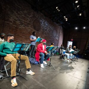 Eight Black artists, Michael-Lamont Lytle, Danté Prince, Michael Clarke, Travae Williams, Rose-Mary Harbans, Krystle Chance, Germaine Konji, and Shakura S'Aida sit or stand on a black stage in front of a brick wall. Each looks at a script set on a music stand in front of them.