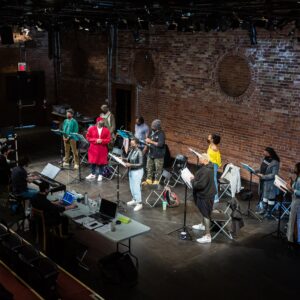 The cast of Dixon Road, Danté Prince, Travae Williams, Michael-Lamont Lytle, Michael Clarke, Germaine Konji, Aadin Church, Rose-Mary Harbans, Shakura S'Aida, Starr Domingue, and Krystle Chance, stand on the stage of the rehearsal hall behind music stands. In front of them, Farnoosh Talebpour, Kat Chin, Haneul Yi, and Jonathan Corkal-Astorga sit behind two tables with open scripts and laptops. Between the tables, Chris Barillaro is playing a keyboard.