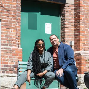 Fatuma Adar and Ray Hogg smile in the sun, sitting on a green bench in front of green doors and a sign that reads “Rehearsal Hall”. Fatuma, a Black woman with long black locs to her hips, wears black sunglasses, a grey-green jumpsuit, and black leather boots. Ray, a Black man with close cropped black hair & a goatee, wears an orange shirt under a dark blue button-down and jeans.