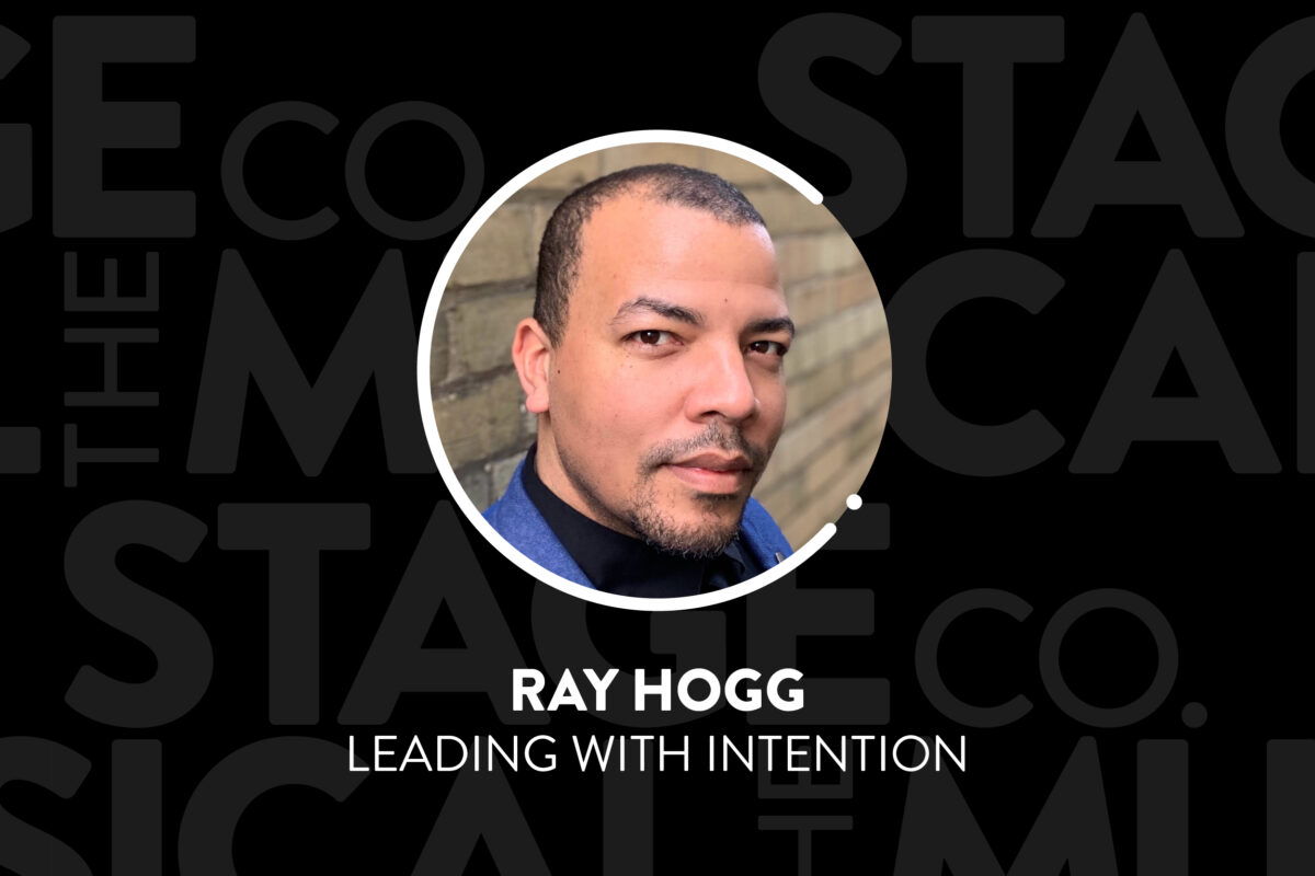 A black background has faded Musical Stage Company logos overlaid. Centered is a circular image of a headshot, with a white 'C' and '.' border. The headshot is Ray Hogg, a Black man, in three quarter profile looking directly into the camera with a slight smile and warmth in his eyes. He has close cropped balck hair, a widow's peak, dark eyebrows and a goatee. He is wearing a blue blazer, a black dress shirt and stands against a yellow brick background. Text underneath reads, “Ray Hogg / Leading with Intention.”