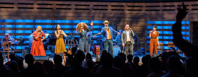 A centered, distanced image of 6 performers lined up and spotlighted on stage, as they sing into their mic and encourage the standing audience to clap along and dance. Left to right shown are Kelly Holiff, a while woman with long blonde hair and green eyes, wearing an orange southern belle dress and cowboy boots; Hailey Gillis, a while woman with long wavy brown hair half-tucked back, wearing a mustard cocktail dress; Jully Black, a Black woman of Jamaican-Canadian descent with long, full earth-toned hair and blonde highlights, wearing a belted navy wrap dress; Beau Dixon, a Black man with warm eyes and a moustache, wearing a blue blazer and light trilby hat; Andrew Penner, a white man with brown and silver hair and a grey blazer, & Sara Farb, a white woman with short, brown curly hair, wearing a belted, clay-colour jumpsuit. Behind them are glimpses of the orchestra and several instruments. Several rows of audience members run along the bottom frame, the theatre darkened.