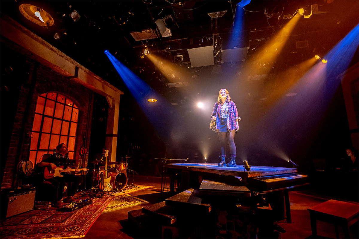 Image of performer Annika Tupper standing on a platform on stage with blue and golden lighting. To the left of her, performer Brandon McGibbon sits holding an electric guitar, surrounded by other band instruments on multiple rugs and a large window and brick wall behind him.