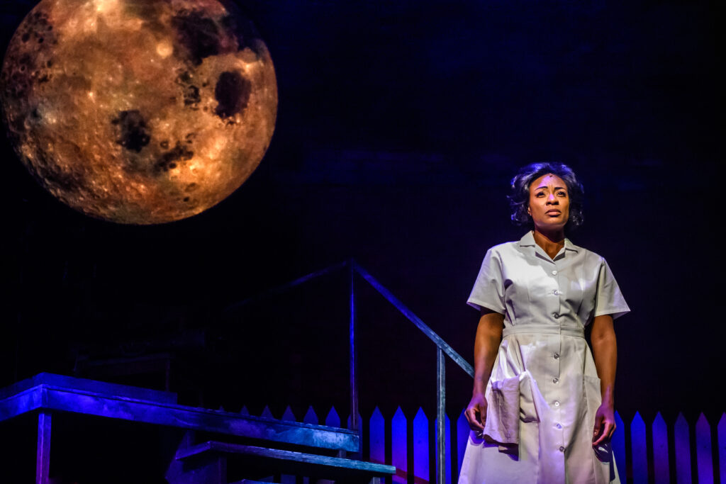 Production photo of Jully Black performing in CAROLINE OR CHANGE. Jully is wearing a maid's uniform, standing in front of a fence, and a platform with two stairs amd a railing. A projection of a moon is on the backdrop behind her.