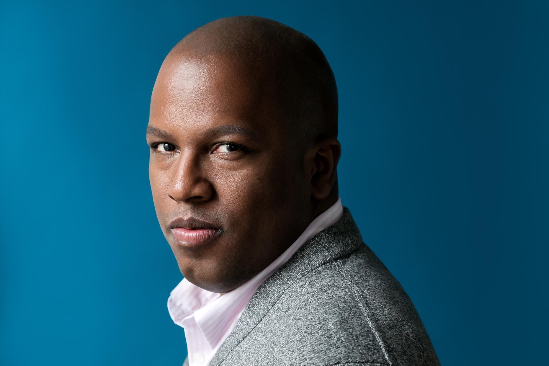 Headshot image of Michael R. Jackson with a blue background. Michael is wearing a collared shirt under a soft grey sweater.