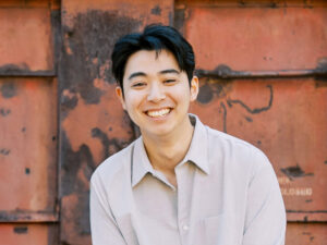Headshot of Kalos Chu. Kalos has black hair and brown eyes, wears a tan long-sleeve shirt against an orange backdrop, and is smiling and looking at the camera.