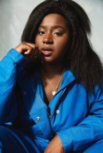 Headshot of Susan Wokoma. Susan is looking at the camera with a hand resting against her cheek. She is wearing a bright blue jacket and gold necklaces.